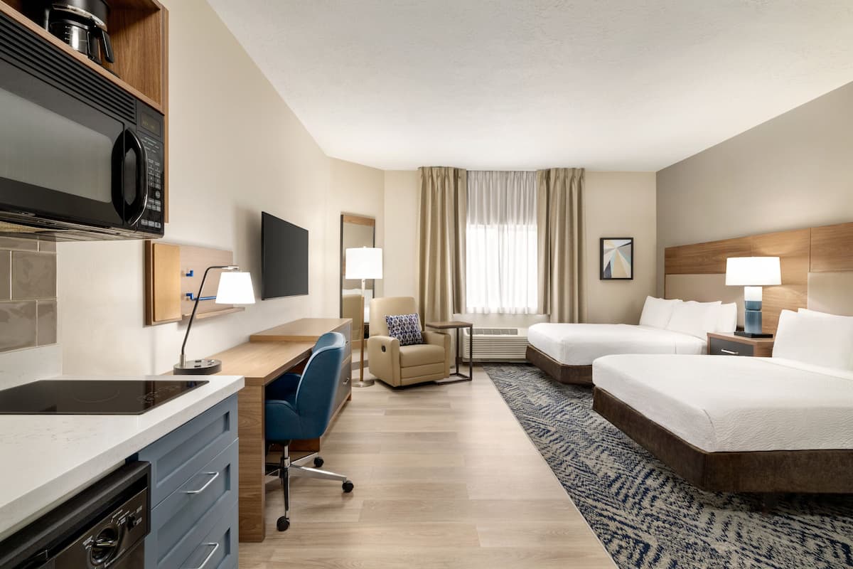 A hotel room with a kitchenette, a desk, a chair, two beds, a TV, and a window with curtains.