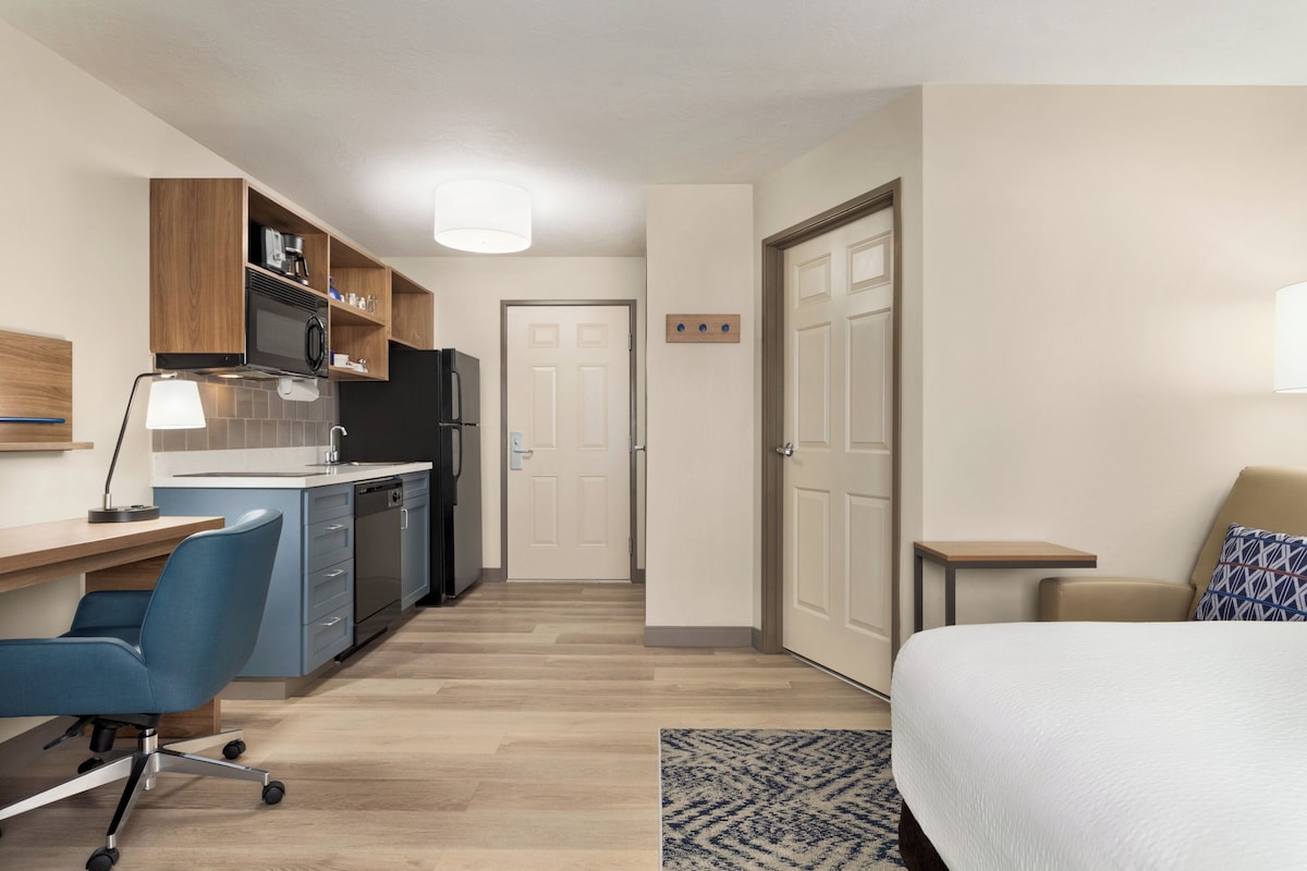 A compact hotel room with a kitchenette, a desk, a chair, a bed, and a small table.