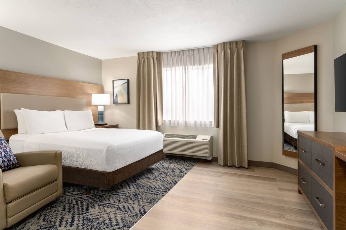 A hotel room with a large bed, a bedside table with a lamp, a dresser, a mirror, and a window with curtains.