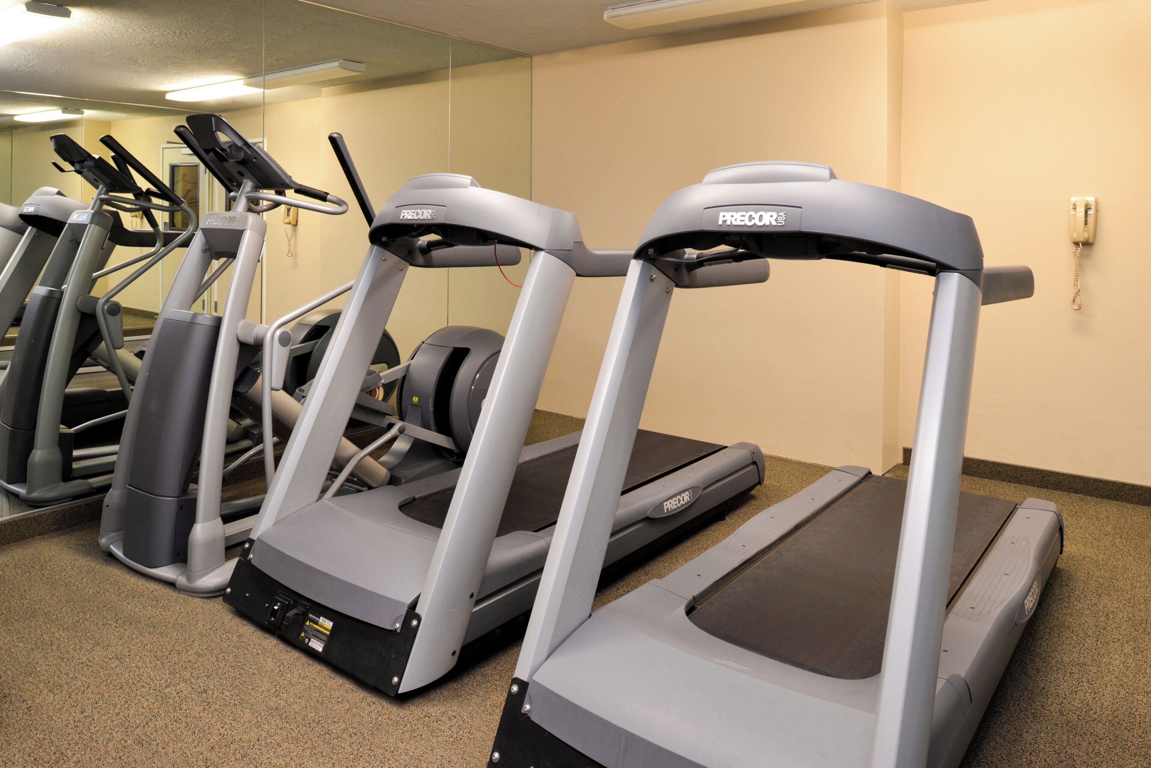 A row of treadmills in a gym with a beige wall and carpeted floor.