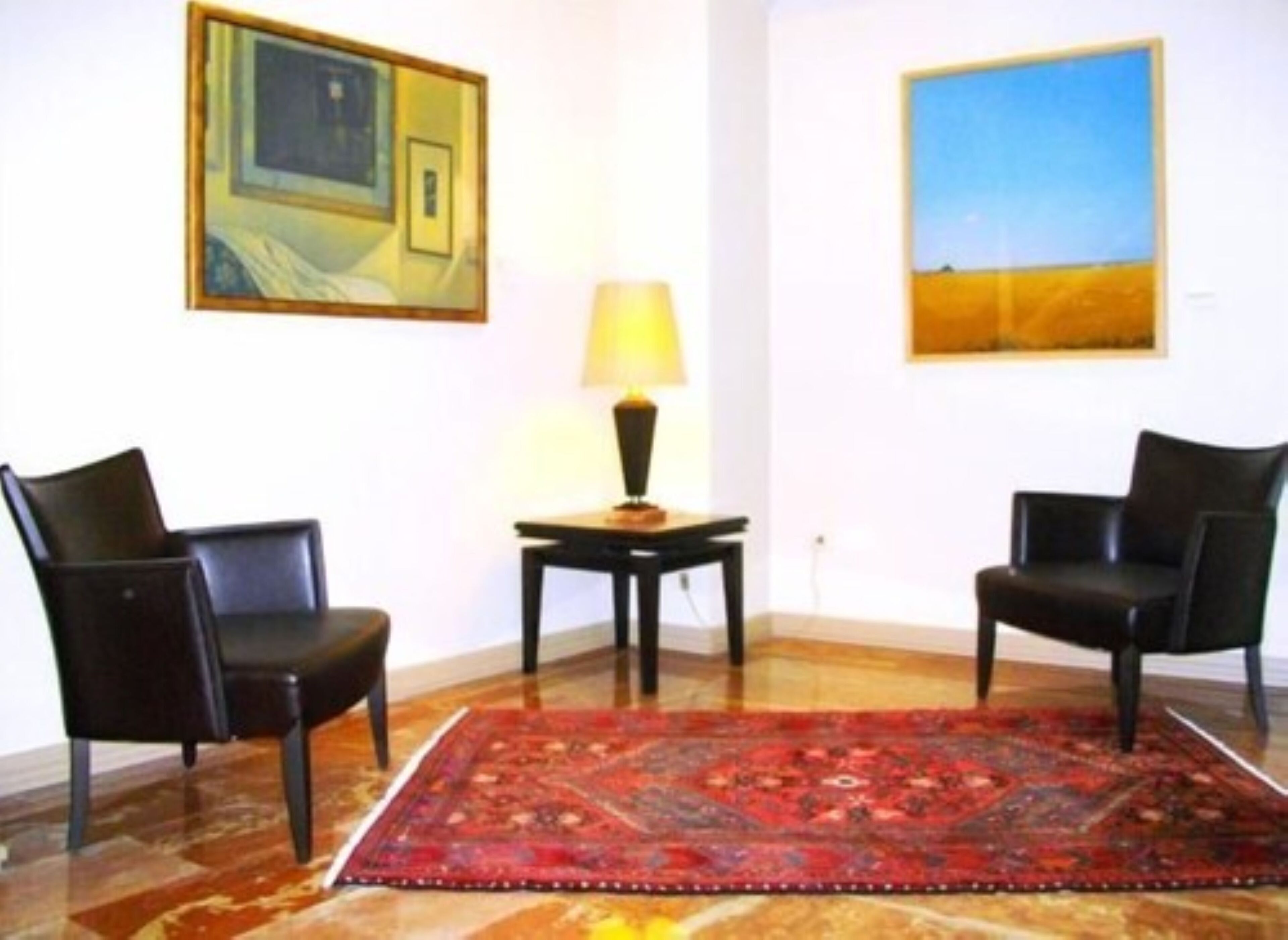 A room with two black leather chairs, a red patterned rug, a table with a lamp, and two paintings on the walls.