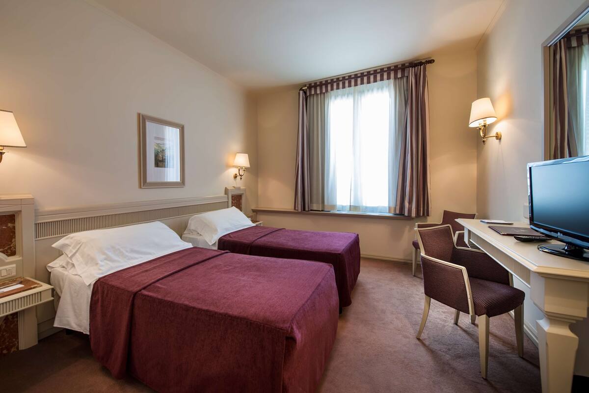 A hotel room with two beds, a desk, a chair, a television, and a window with curtains.