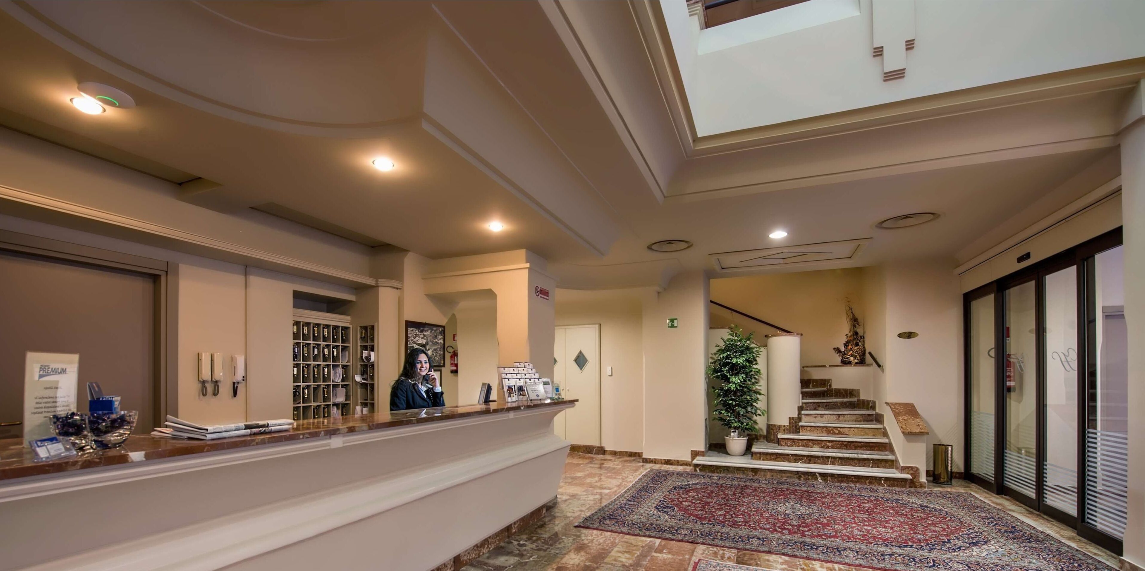 A hotel reception area with a reception desk, a staircase, and a large window.