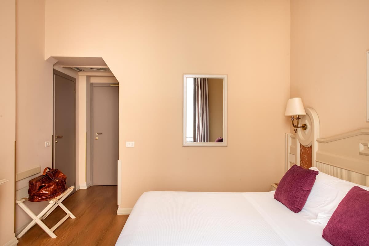 A hotel room with a bed, a nightstand, a lamp, a mirror, and a bench with a handbag.