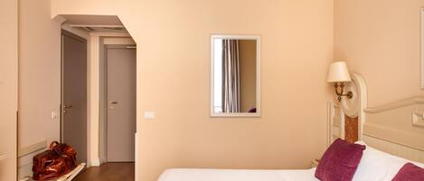 Una habitación de hotel con cama, mesita de noche, lámpara, espejo y un banco con un bolso de mano.