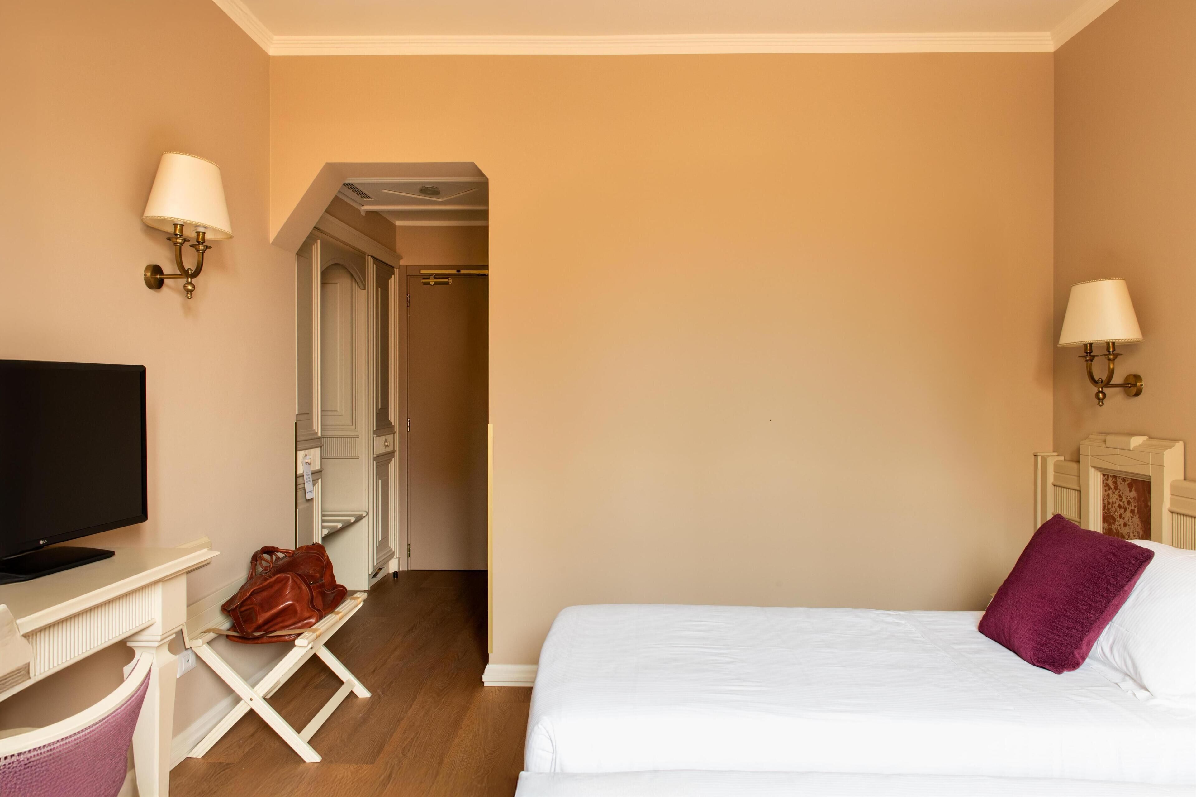 Una camera d'albergo con un letto, una scrivania con televisore, una sedia, una borsa e una porta che dà su un'altra stanza.