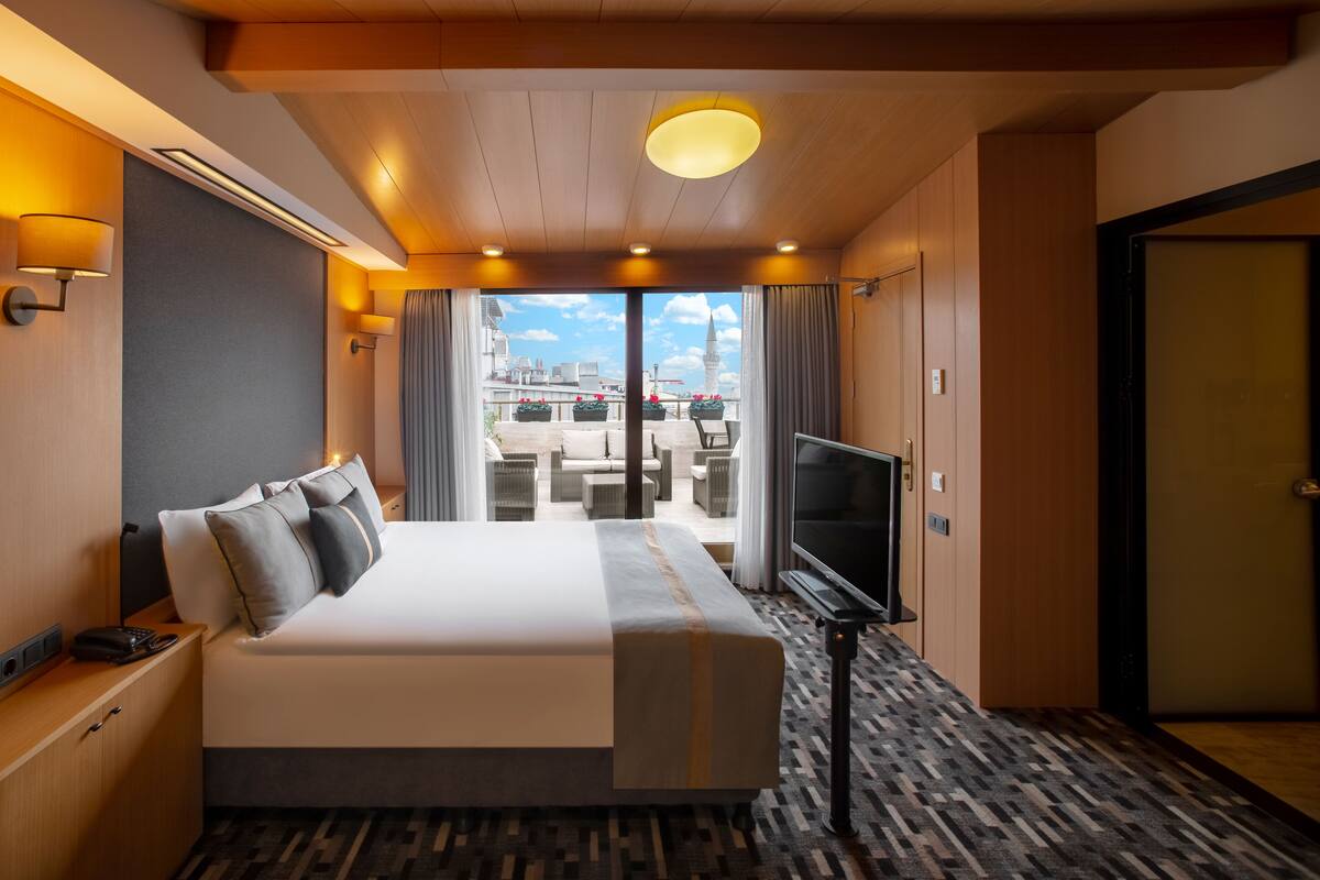 A hotel room with a large bed, a TV, a balcony with outdoor seating, and a city view.