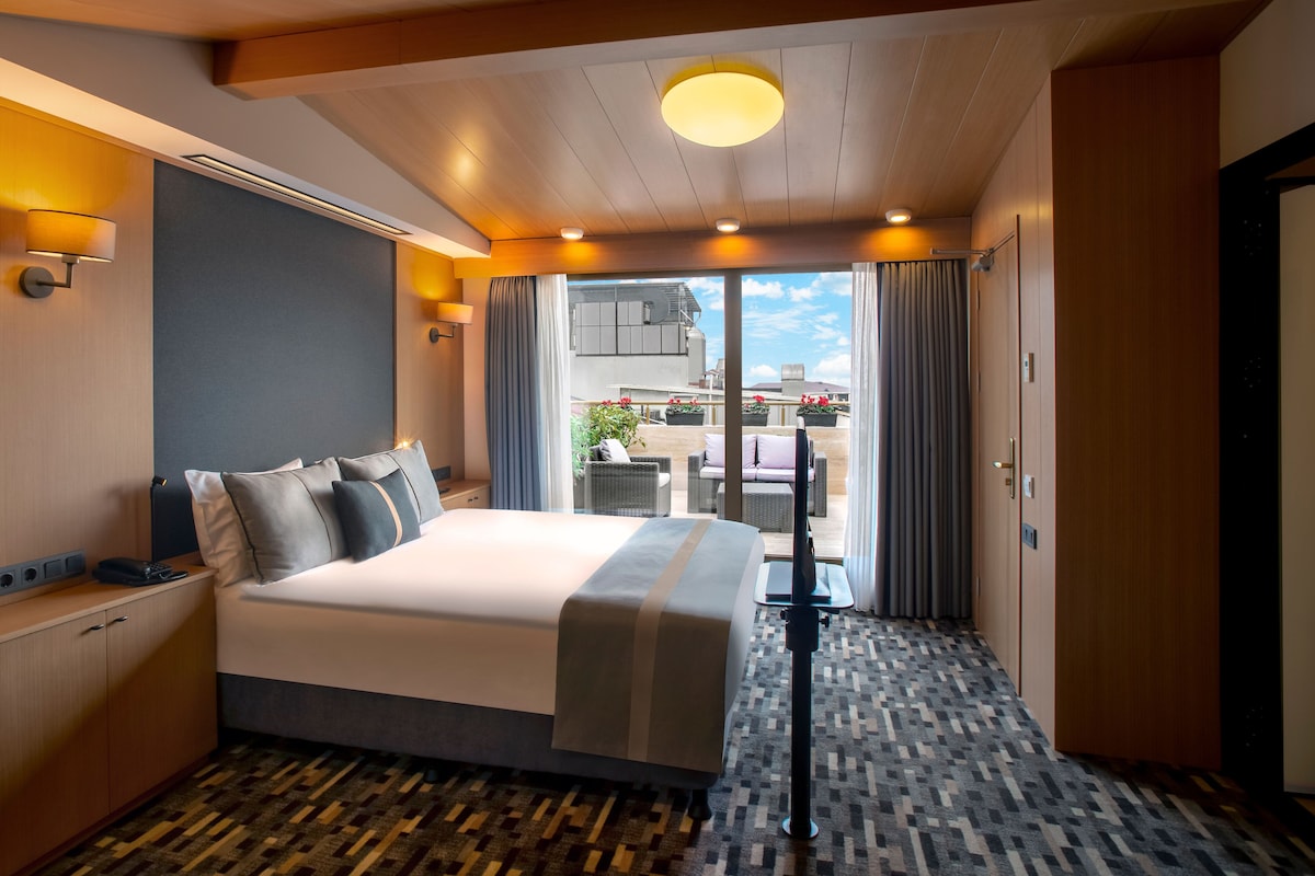 Habitación de hotel con una cama grande, un escritorio con computadora, vista al balcón exterior y una lámpara del techo.