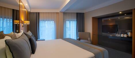 A modern hotel room with a large bed, a seating area, a television, and a kitchenette.