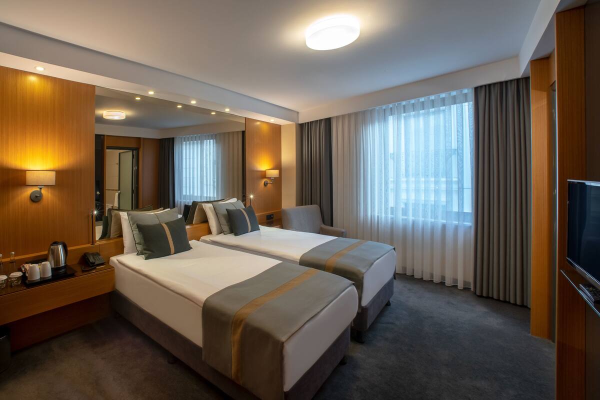 A hotel room with two beds, a flat-screen TV, a minibar, and a large window with curtains.