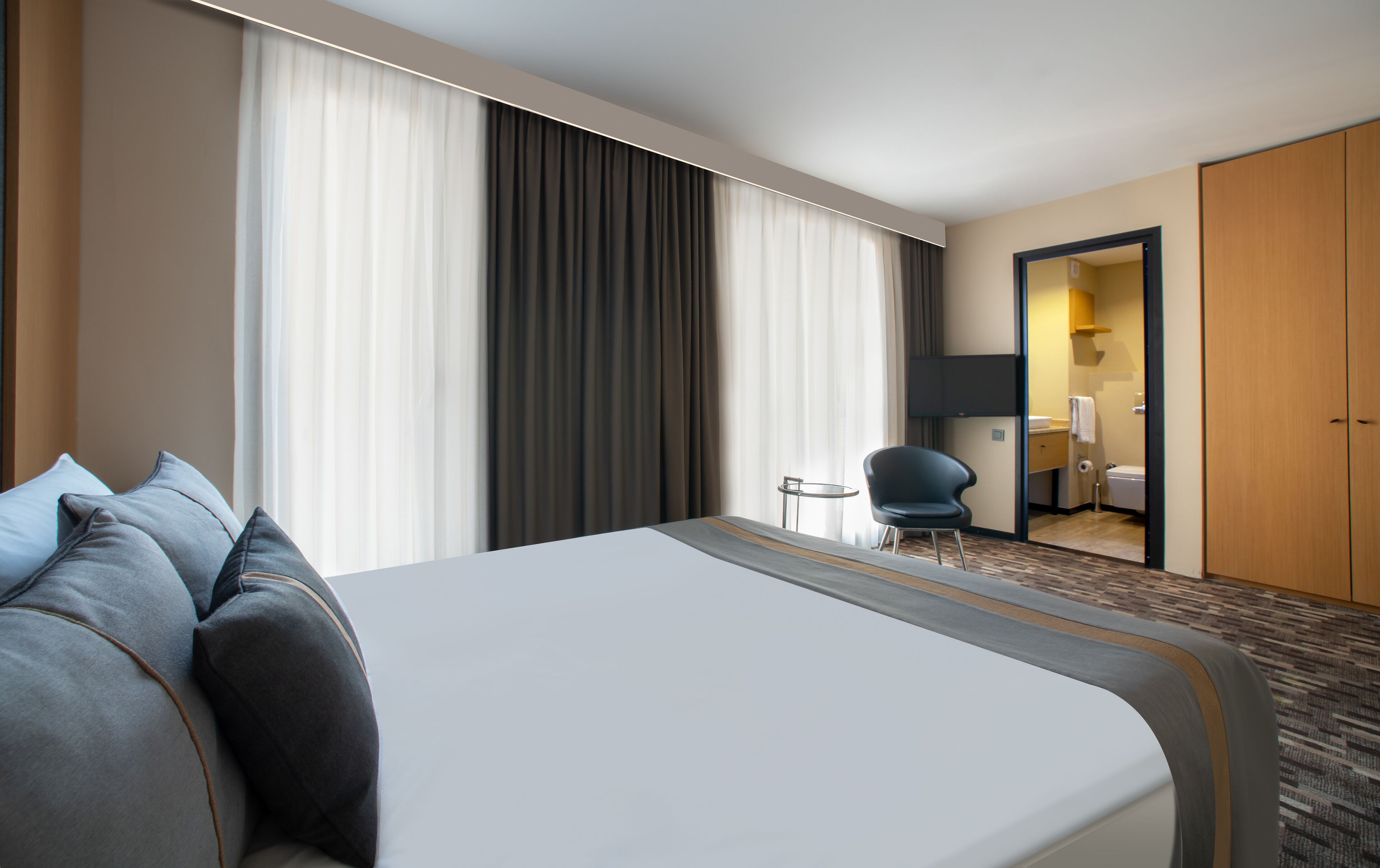 Quarto de hotel com uma cama grande, uma cadeira, uma mesinha e um banheiro visível através de uma porta aberta.