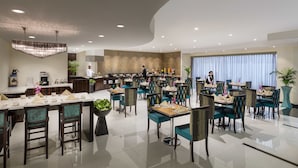 A modern dining area with multiple tables and chairs, a kitchen in the background, and a large window with curtains.