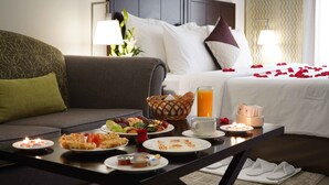 A hotel room with a dining table set with breakfast items, a bed with rose petals, and a lamp.