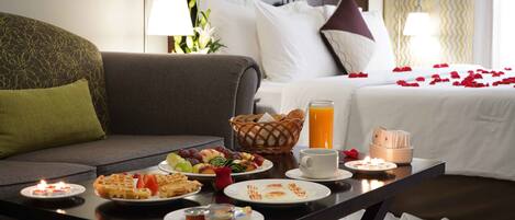 A hotel room with a dining table set with breakfast items, a bed with rose petals, and a lamp.