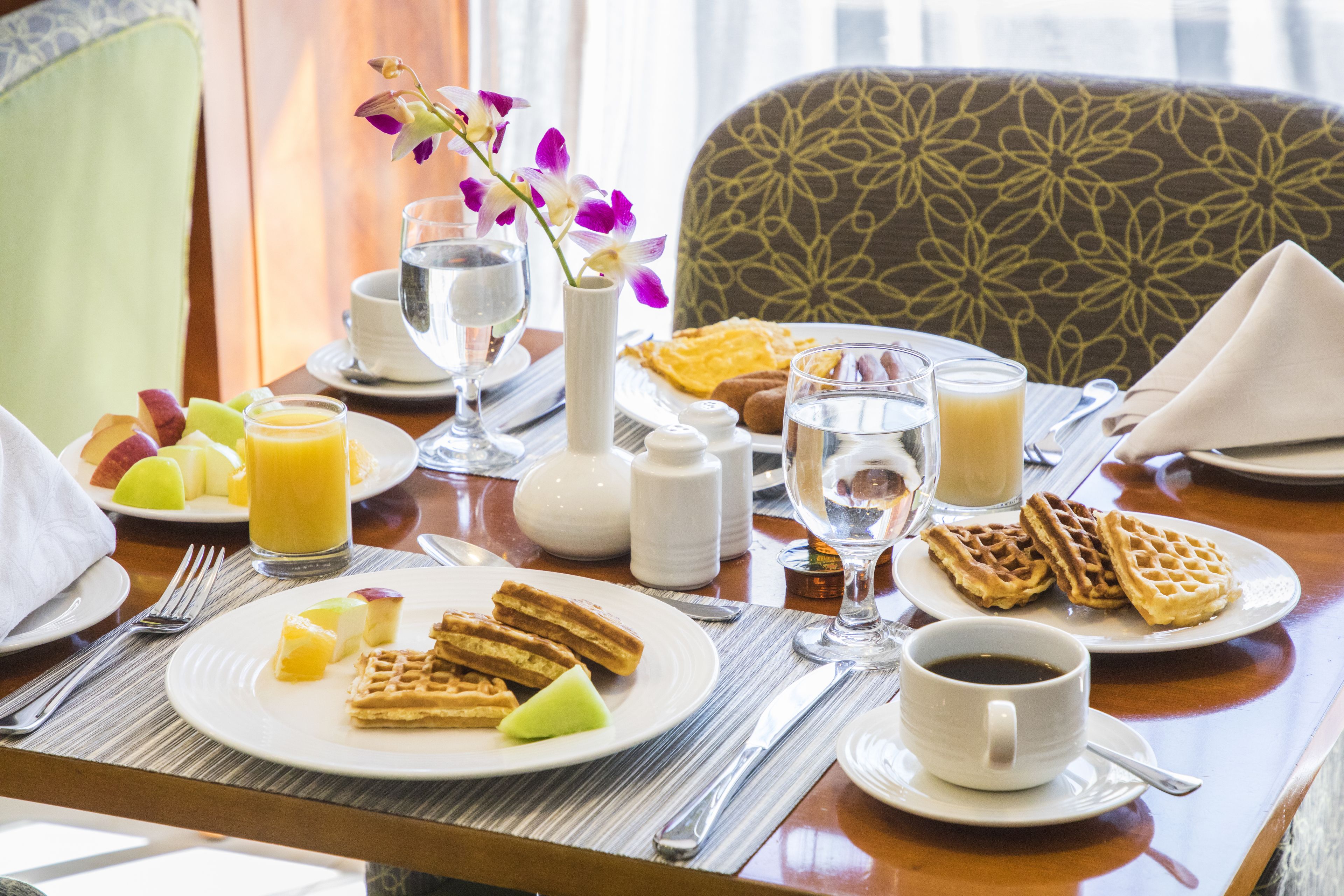 A breakfast setting with plates of waffles, fruit, and juice, a vase with flowers, and a cup of coffee on a table.