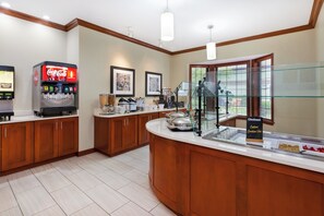 A hotel breakfast area with a Coca-Cola vending machine, a buffet counter with a sign for Chobani, and framed artwork on the wall. - Staybridge Suites Kalamazoo by IHG (Kalamazoo)