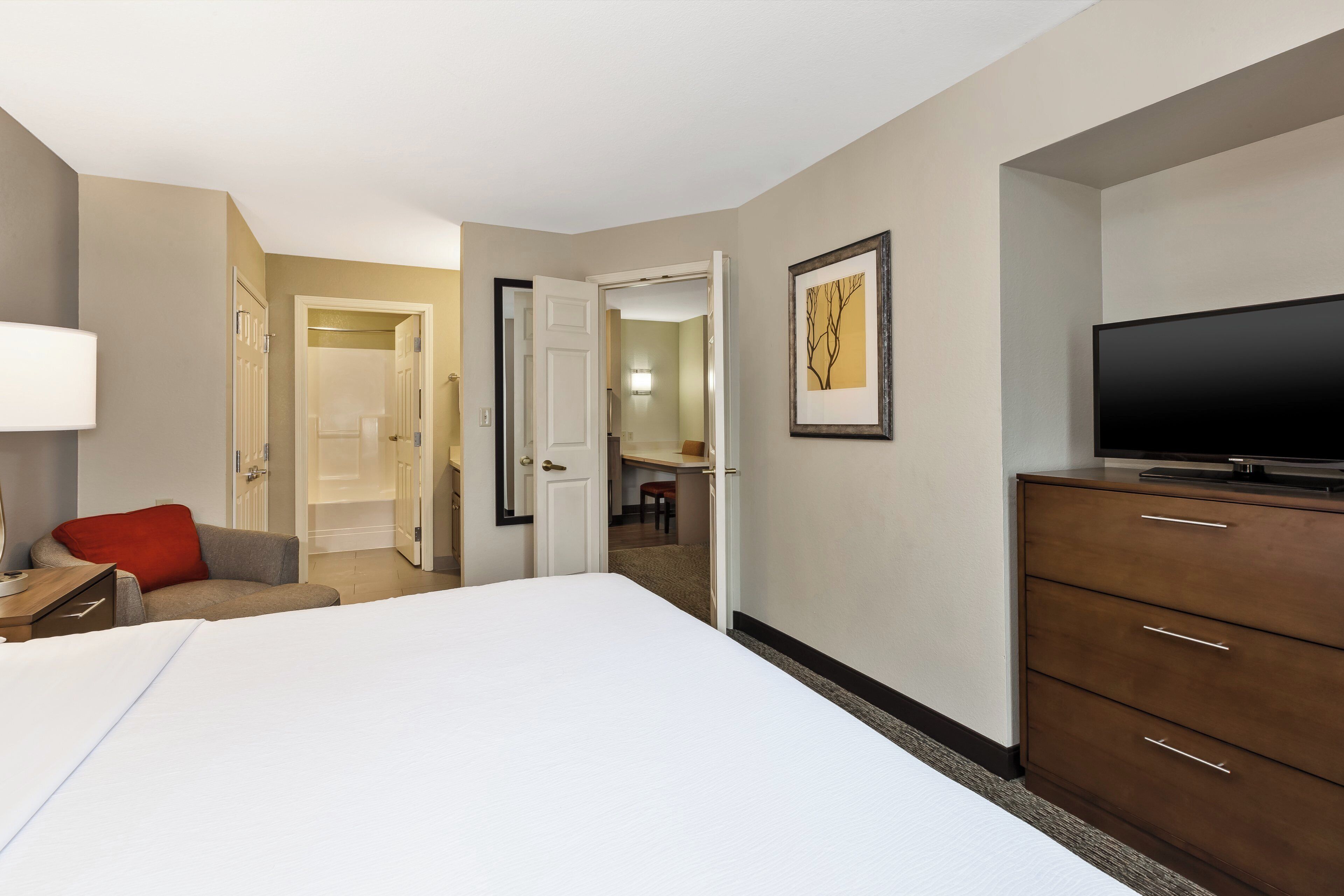 A hotel room with a bed, a dresser, a television, a desk, and a bathroom visible through an open door.