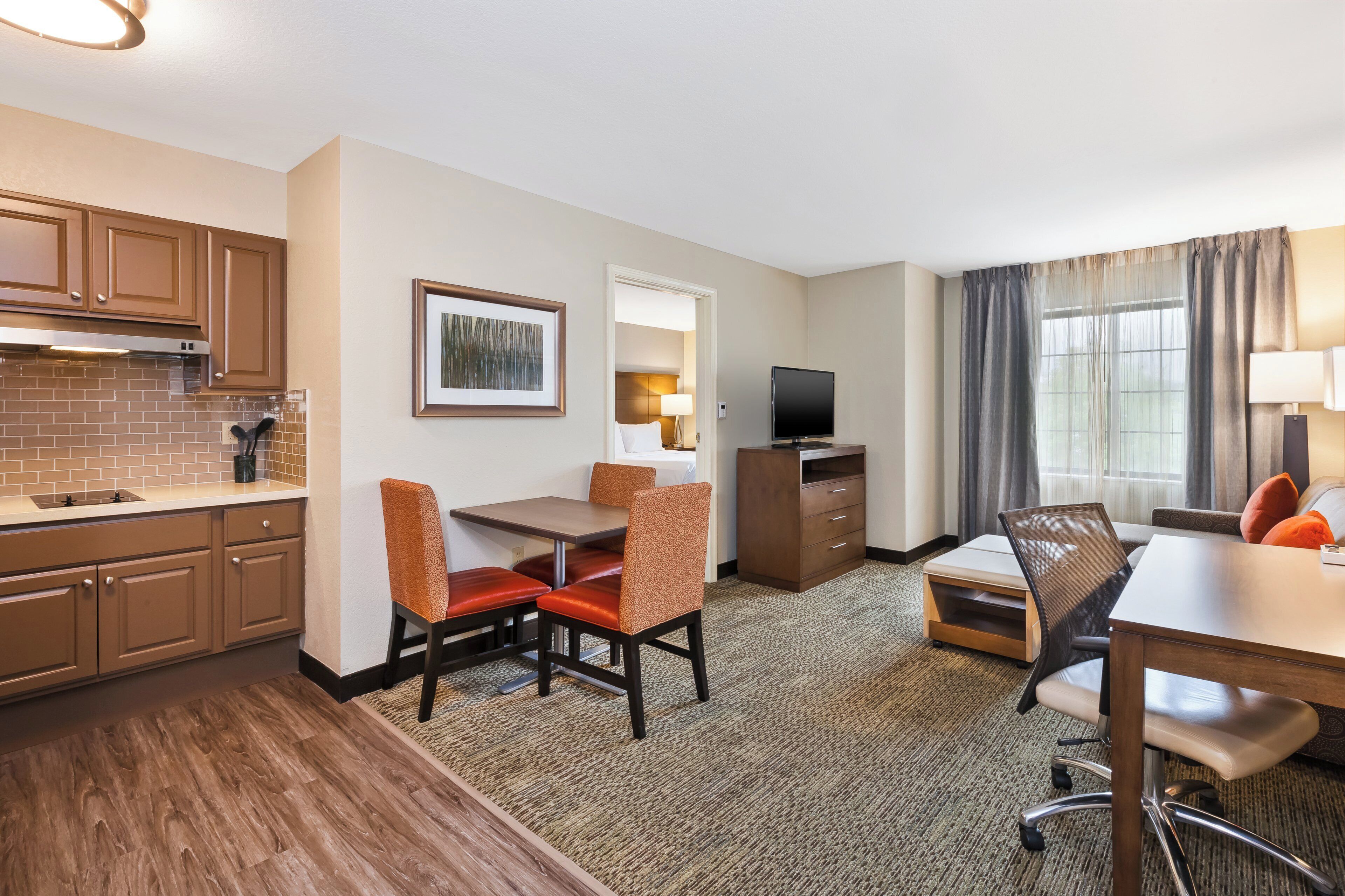 Een hotelkamer met een kitchenette, een eethoek met stoelen en een tafel, een televisie en een raam met gordijnen.
