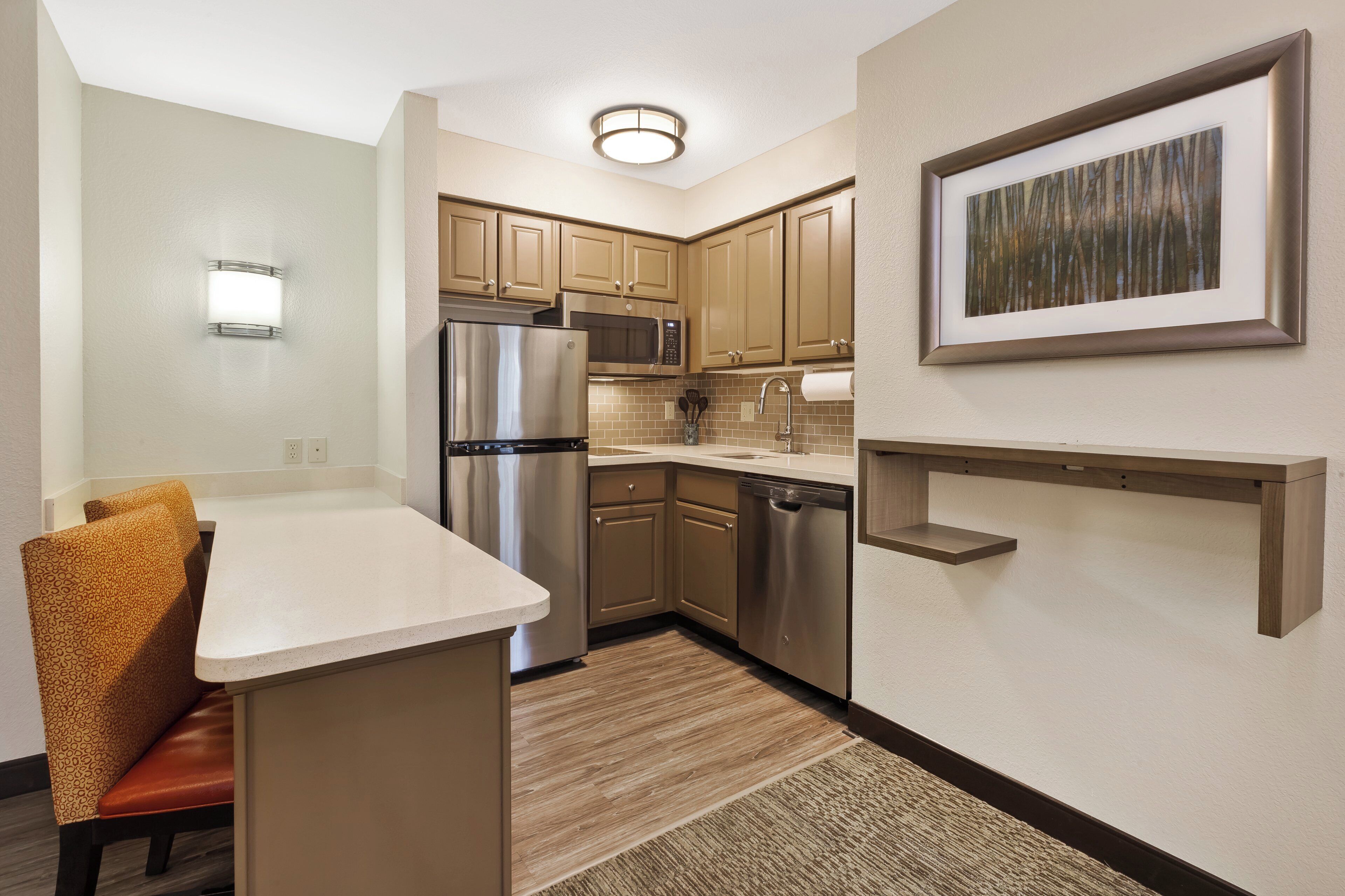 A compact kitchen with a refrigerator, microwave, and dishwasher, a small dining area with a chair, and a framed picture on the wall.