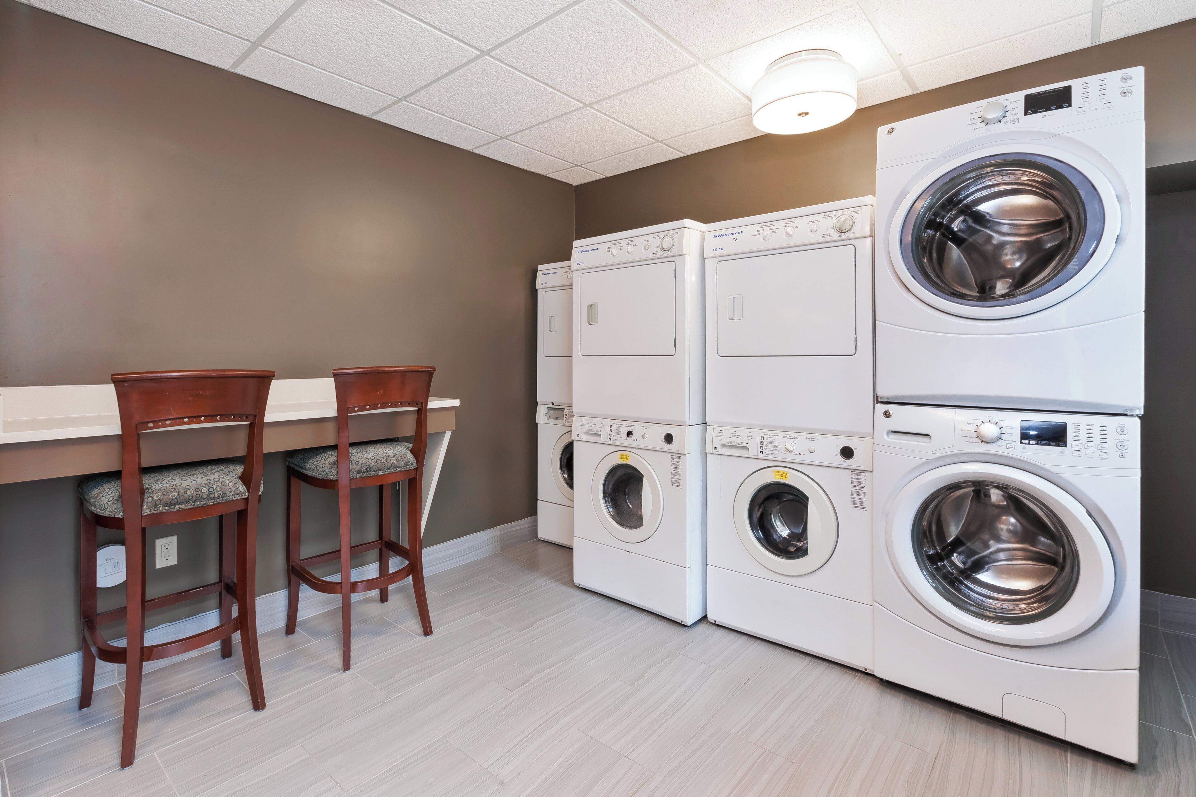 A laundry room with stacked washers and dryers, wooden chairs, and a light fixture on the ceiling.