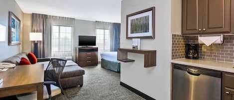 Ein Hotelzimmer mit Bett, Schreibtisch mit Stuhl, Fernseher, Küchenzeile inklusive Kaffeemaschine und einem gerahmten Bild an der Wand.