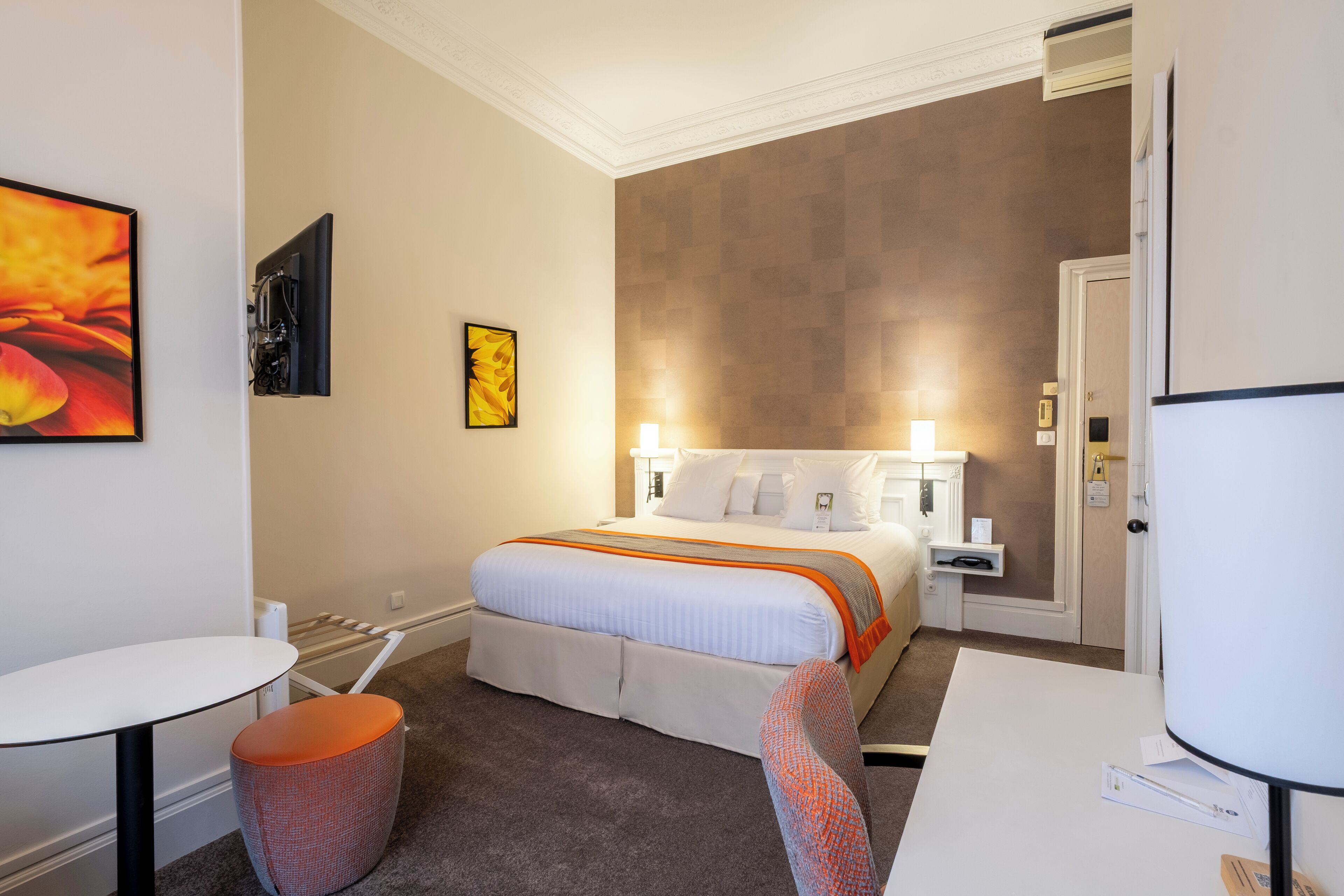 A hotel room with a bed, a desk, a chair, a small table, and artwork on the walls.