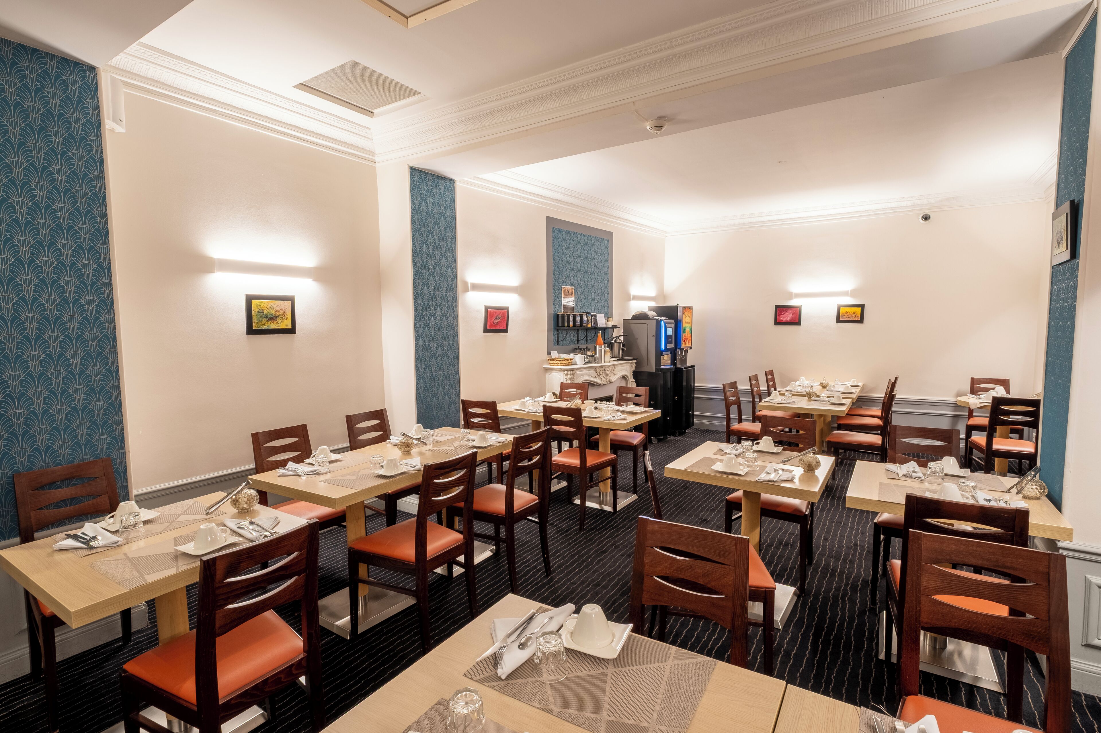 A hotel dining area with multiple tables and chairs, light fixtures on the walls, and a refrigerator in the background.