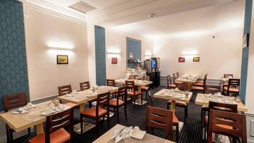 A hotel dining area with multiple tables and chairs, light fixtures on the walls, and a refrigerator in the background.