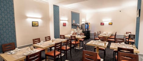 A hotel dining area with multiple tables and chairs, light fixtures on the walls, and a refrigerator in the background.