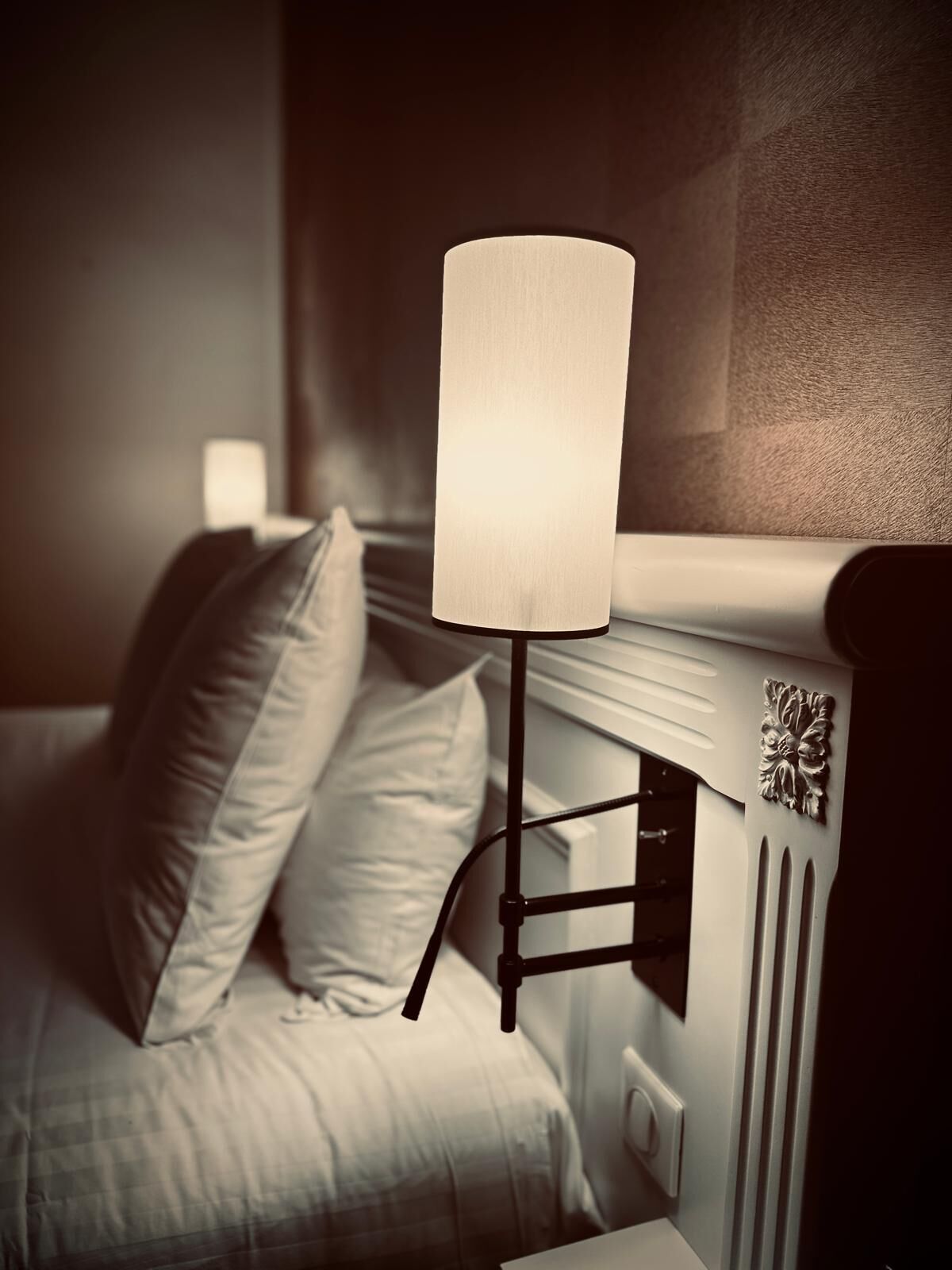 Un letto con copriletto bianco e due cuscini, un comodino con una lampada e un elemento decorativo a parete.
