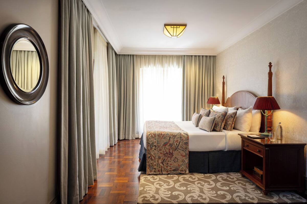 A hotel room with a bed, bedside lamps, a mirror, curtains, a patterned rug, and a wooden nightstand.