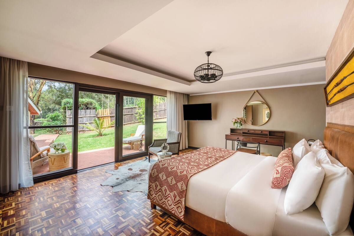 A hotel room with a large bed, a TV, a wooden dresser, and a sliding glass door leading to a garden.