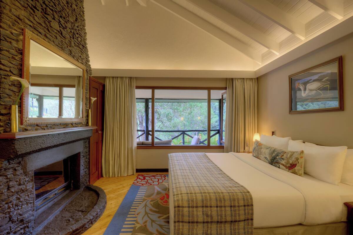 A hotel room with a stone fireplace, a large bed with white linens, a painting on the wall, and a view of the outdoors through large windows.