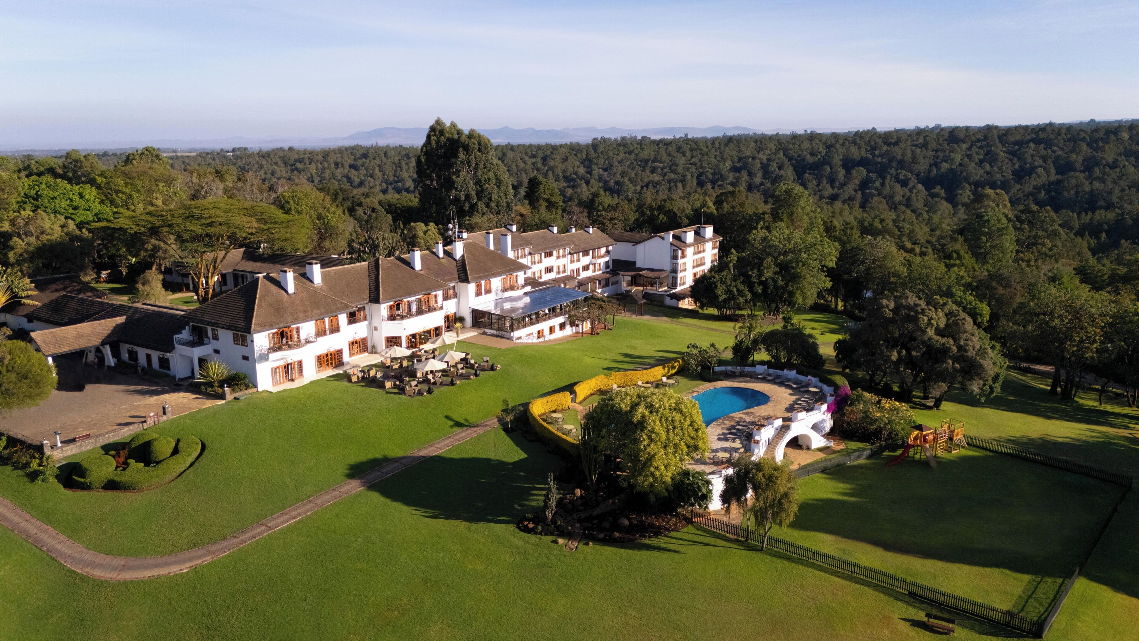 A large, multi-story hotel with a well-maintained lawn, a swimming pool, and a tennis court.