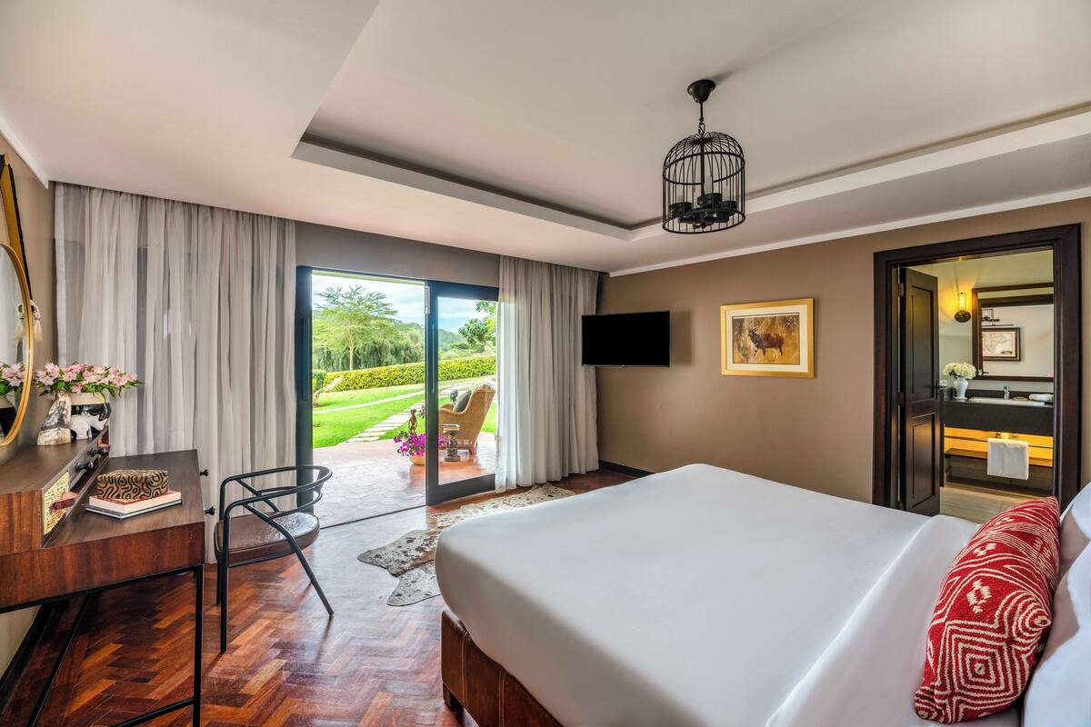 A hotel room with a large bed, a desk with a chair, a TV mounted on the wall, and a sliding glass door leading to a garden.