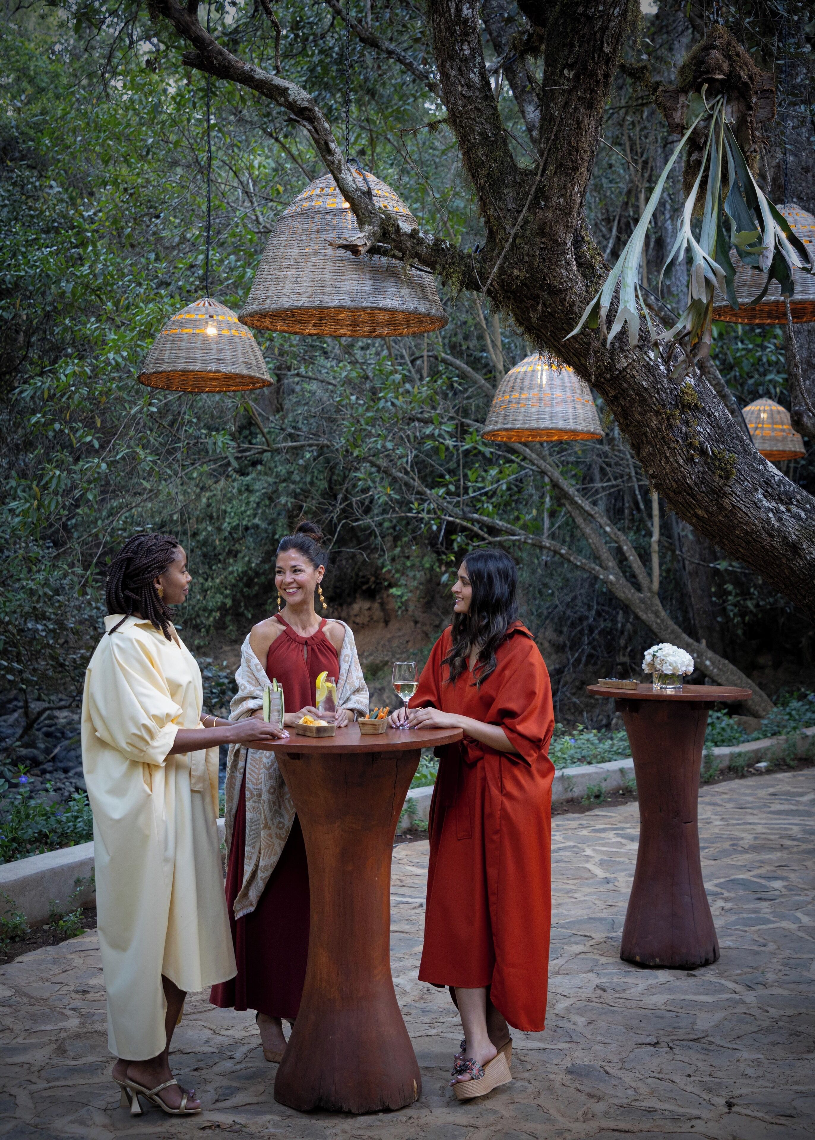 Three people standing around a round wooden table with food and drinks, surrounded by trees and hanging wicker lamps.