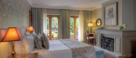 A hotel room with a large bed, a fireplace, a desk with a lamp, and a view of the outdoors through large windows.