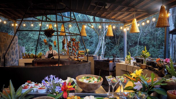 An outdoor dining area with a chef preparing food, surrounded by hanging lights and a view of the forest. - Fairmont Mount Kenya Safari (Nanyuki)