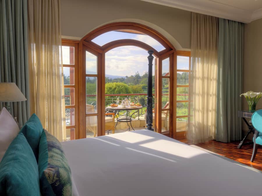 A bedroom with a large bed, a view of a balcony with a table and chairs, and a chair with a vase of flowers.