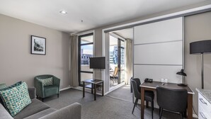 A hotel room with a grey sofa, a green armchair, a desk with a computer, a TV, and a large window offering a city view. - Quest on Queen (Auckland)