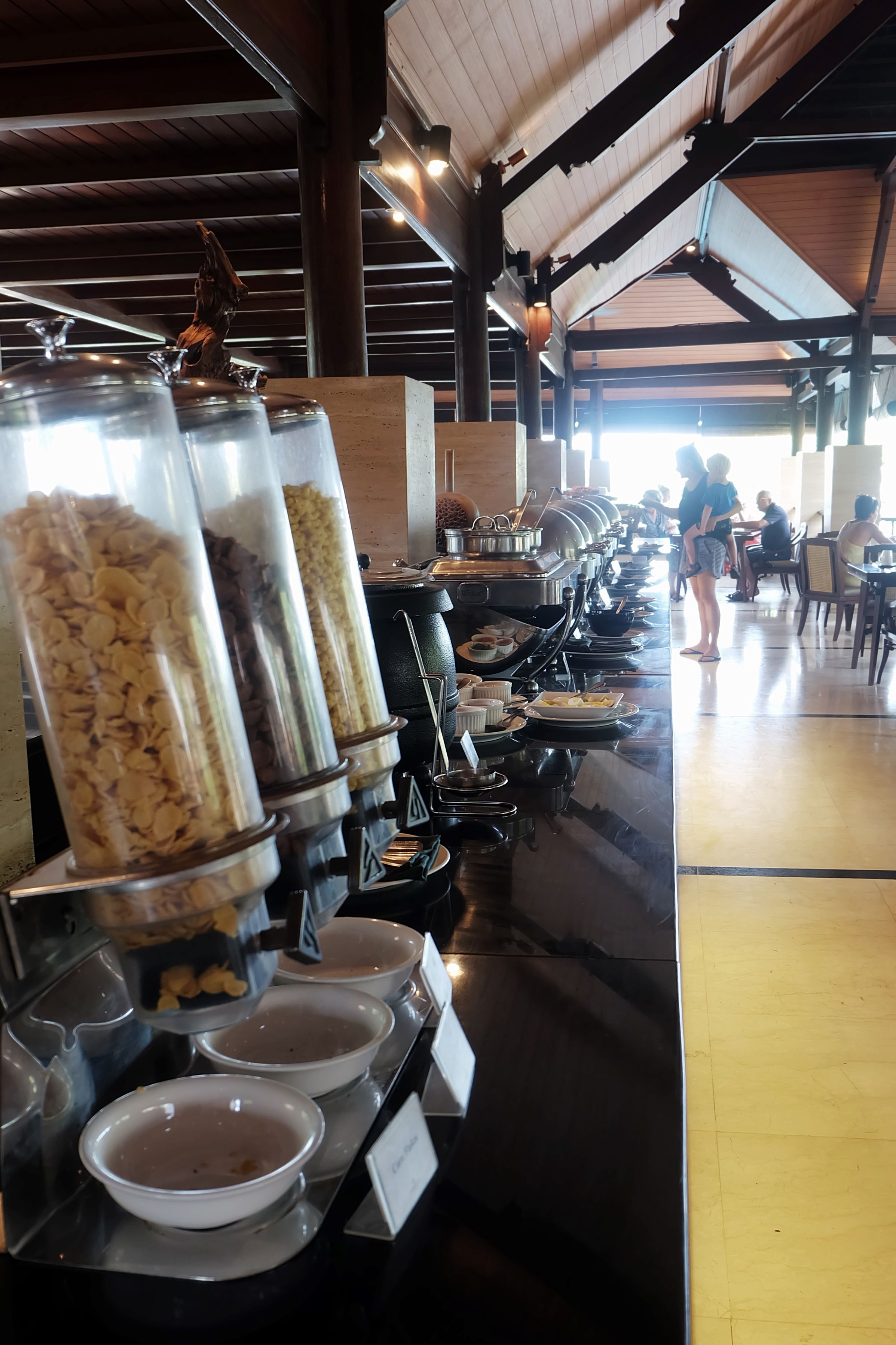 Un buffet avec différents récipients et bols remplis de nourriture, un plafond en bois et une personne debout à l’arrière-plan.