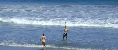 A beach scene with two people, one standing in shallow water and the other walking along the shore.