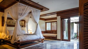 A room with a four-poster bed, a bedside table with a lamp, and a large window with wooden shutters. - Grand Balisani Suites Hotel (Keroboken Kelod)