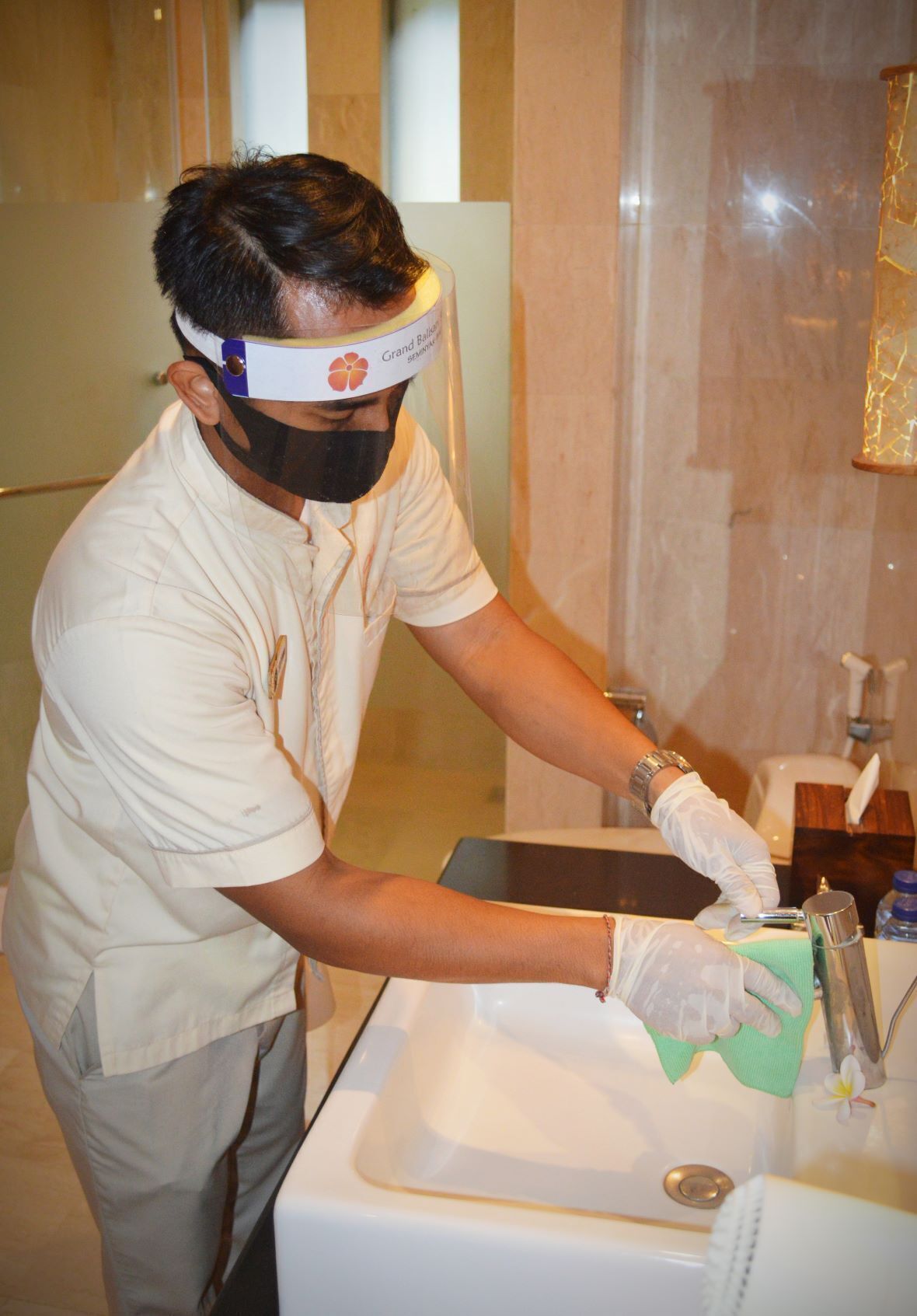 A person cleaning a sink in a bathroom with a marble wall and a glass shower enclosure.