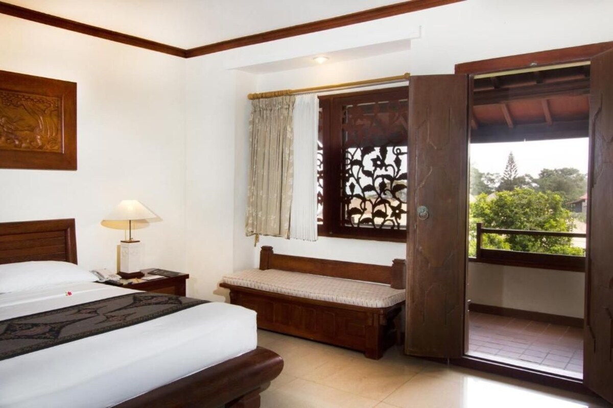 A hotel room with a bed, bedside table, lamp, bench, window with curtains, and a balcony door.