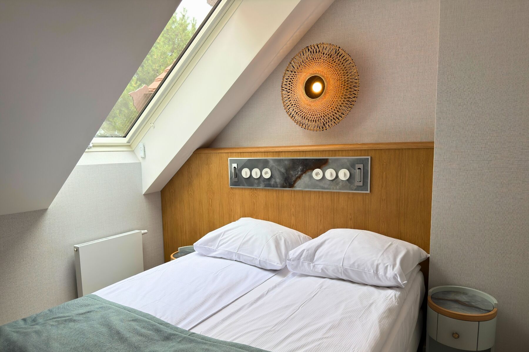 A modern bedroom with a sloped ceiling, a bed with white linens, a wooden headboard, a wall-mounted light fixture, and a window with a view of trees.