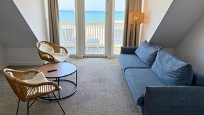Una habitación con un sofá gris, dos sillas de mimbre, una mesita redonda y vista a la playa a través de un ventanal.