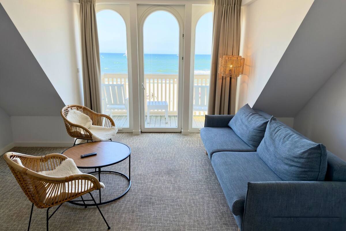 Una habitación con un sofá gris, dos sillas de mimbre, una mesita redonda y vista a la playa a través de un ventanal.