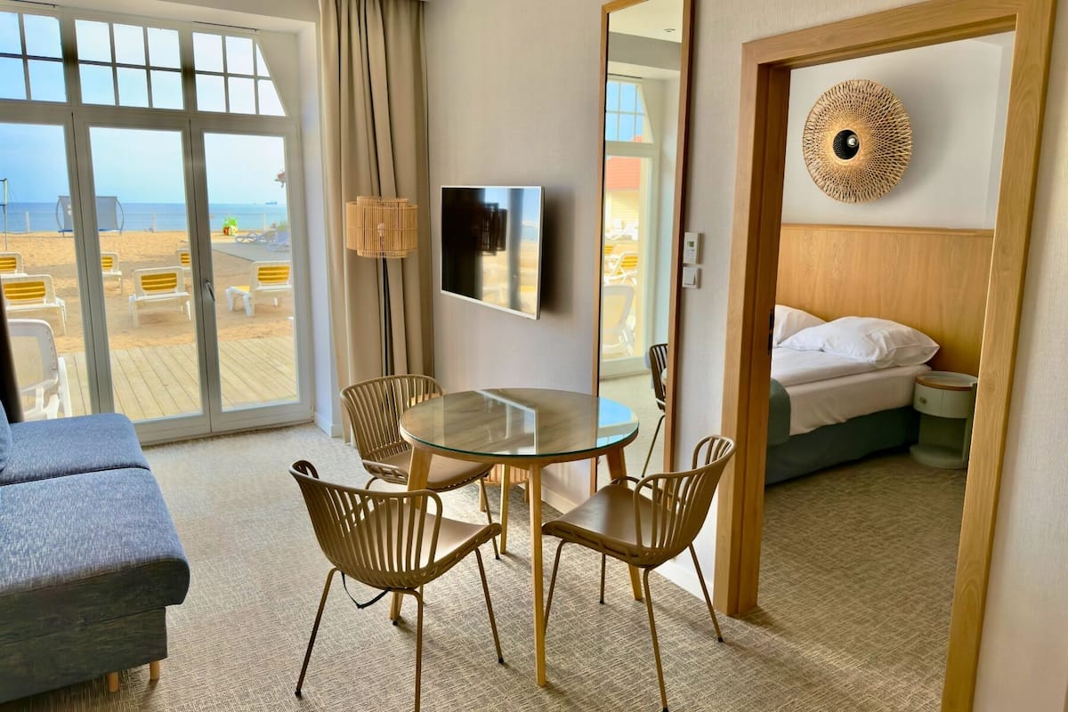 Habitación de hotel con puerta de vidrio que da a vista de playa, una mesa redonda de vidrio con sillas, un televisor en la pared y una cama con ropa blanca.