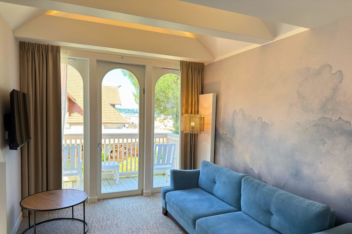 Una sala de estar con un sofá azul, una mesita redonda y un televisor montado en la pared. Hay una puerta corrediza de vidrio que da a un espacio exterior con una terraza de madera y vistas a edificios.